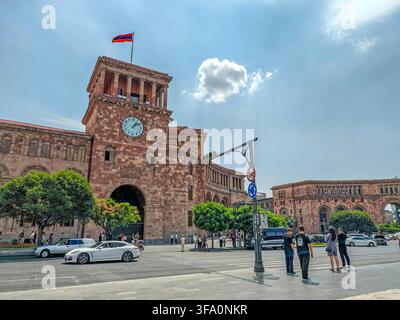 Government House with clock tower building with clock tower and Armenian flag. Cars move near pedestrians on sunny day. Green trees line street beside Stock Photo