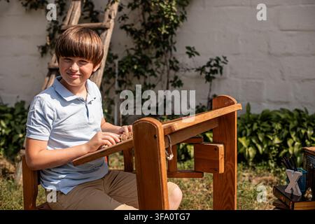 A boy sits at a wooden workbench in a garden. He is focused on his project and enjoys his time outside. The sun shines and plants grow in the backgrou Stock Photo