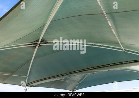 Looking up at the underside of a large, green mesh shade sail structure with white metal supports against a blue sky Stock Photo