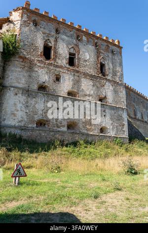 Weathered stone walls of ancient Medzhybizh fortress palace feature arched windows and cracked masonry. Construction warning sign stands on green gras Stock Photo