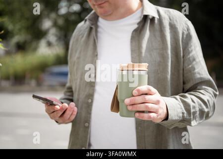 Man holding reusable travel mug and smartphone outdoors Stock Photo