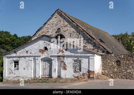 Ancient historical building features peeling white plaster, exposed brickwork, and closed wooden door. Weathered masonry structure stands under clear Stock Photo