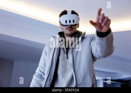 Young man wearing a virtual reality headset reaches forward with his hand while standing in a contemporary indoor space, exploring immersive digital t Stock Photo