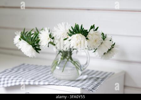 Delicate white flowers and green foliage arranged in a clear glass pitcher, resting on a black-and-white checkered cloth against a softly lit white wo Stock Photo