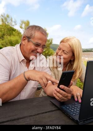 Smiling senior couple sit together at a beach cafe, using a smartphone and laptop. They enjoy digital connection, travel and relaxed retirement lifest Stock Photo