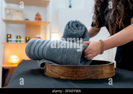 Close-up of a therapist arranging rolled gray towels in a wooden bowl before a relaxing massage treatment at a modern wellness clinic with warm ambien Stock Photo