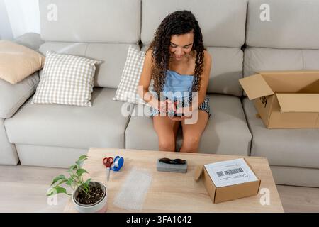 Young woman uses a smartphone while preparing a sold second-hand item for shipping, with cardboard boxes, bubble wrap, scissors, and a parcel label at Stock Photo