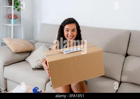 Smiling young woman holds a sealed cardboard box with shipping label at home, preparing a sold item for delivery through an online second-hand marketp Stock Photo
