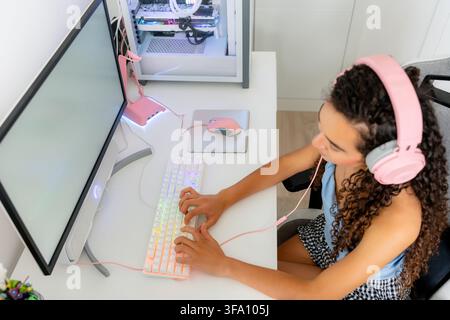 Young woman wearing pink headphones plays video games on a desktop computer with RGB keyboard and gaming mouse at a bright home workstation. Stock Photo