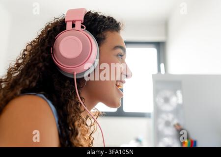 Side view of a smiling young woman wearing pink headphones while playing a video game in a bright gaming room, enjoying online entertainment and immer Stock Photo