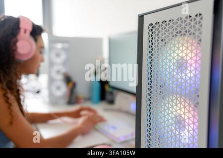 Young woman wearing pink headphones plays video games on a desktop computer in a modern home setup, with colorful RGB lighting visible through the gam Stock Photo