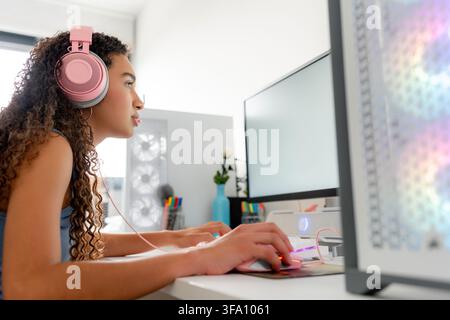 Young woman wearing pink headphones plays a computer game at a desktop workstation, using a keyboard and mouse beside a monitor in a bright home offic Stock Photo