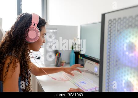 Young woman wearing pink headphones plays a computer game at a desktop setup, using an illuminated keyboard and dual monitors in a bright modern home Stock Photo