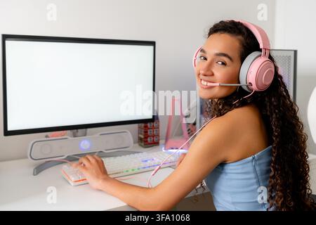 Smiling young woman wearing a pink gaming headset sits at a desktop computer, using a keyboard and mouse beside a blank monitor in a modern home works Stock Photo