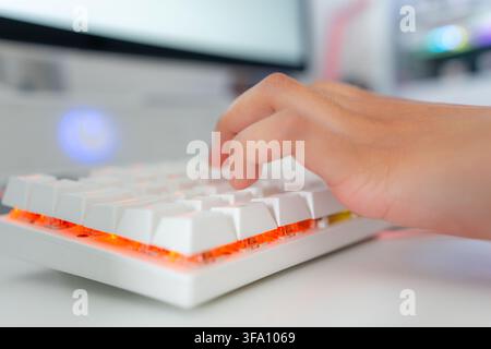 Close-up of a hand pressing keys on a white illuminated mechanical keyboard, capturing a modern gaming room setup with colorful LED lighting and compu Stock Photo