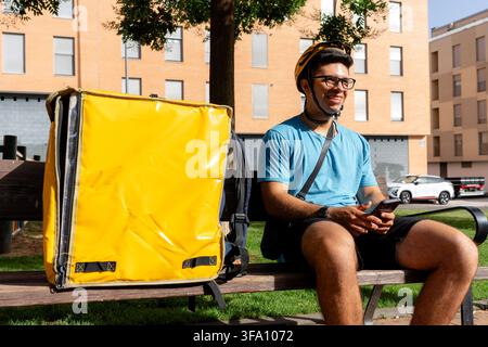 Smiling delivery courier in helmet and blue shirt checks a smartphone while sitting on a park bench beside a large yellow insulated food delivery bag. Stock Photo