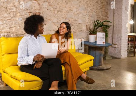 Two female coworkers discuss a printed document while sitting on a bright yellow sofa in a relaxed creative office, illustrating teamwork, communicati Stock Photo