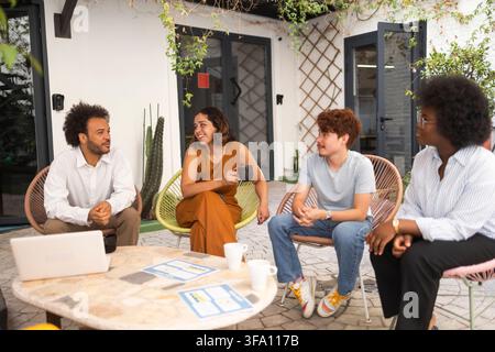 Diverse coworkers gather around a table in a sunny outdoor office, discussing ideas and collaborating during an informal team meeting with a laptop an Stock Photo