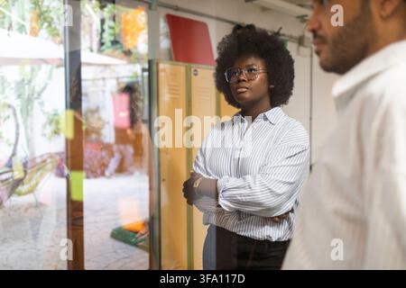 Diverse coworkers discuss ideas in a modern shared office, with a confident woman listening attentively beside a colleague near glass walls and locker Stock Photo