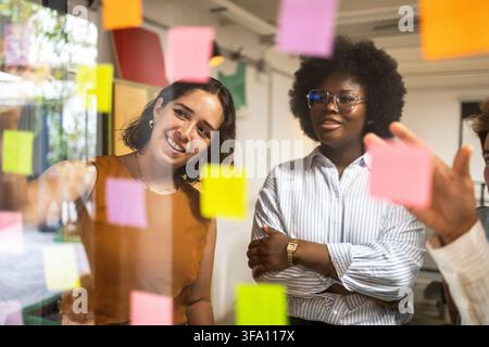 Diverse colleagues review colorful sticky notes on a glass wall during a brainstorming session in a modern coworking office, collaborating on ideas an Stock Photo