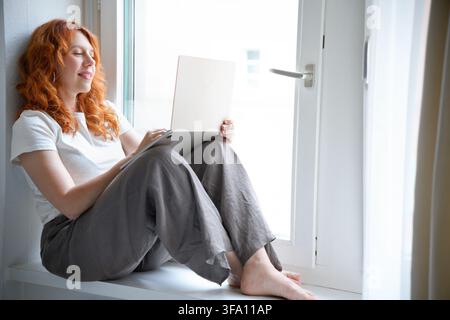 Redhead woman freelancer sits barefoot by a bright window, using a laptop while working remotely from home. Calm digital nomad enjoys a flexible lifes Stock Photo