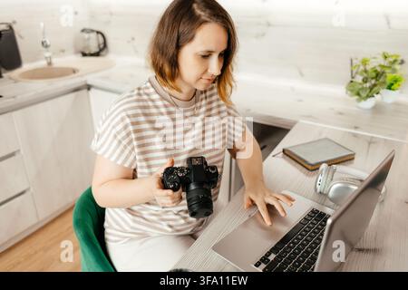 Female photographer sits at a kitchen table with camera in hand, using a laptop to edit images and manage remote work from a bright modern home worksp Stock Photo