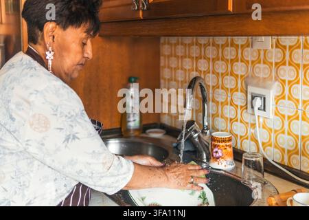 An old woman washes dishes by hand at the kitchen sink in her home, wearing an apron as water runs from the faucet beside patterned tile and wooden ca Stock Photo