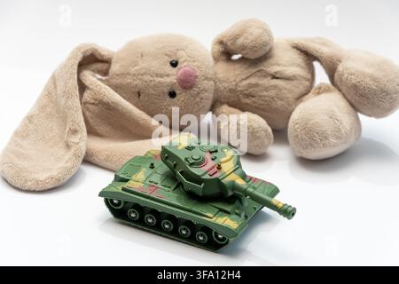 A soft stuffed rabbit lies next to a small green toy tank on a white background. Stock Photo