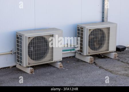 Two beige outdoor air conditioning units are positioned side-by-side on a paved surface against a white corrugated wall Stock Photo