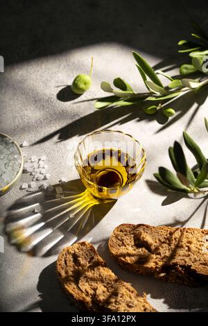 Golden olive oil in a faceted glass bowl beside rustic bread, sea salt, green olive and fresh olive branches, illuminated by dramatic natural sunlight Stock Photo