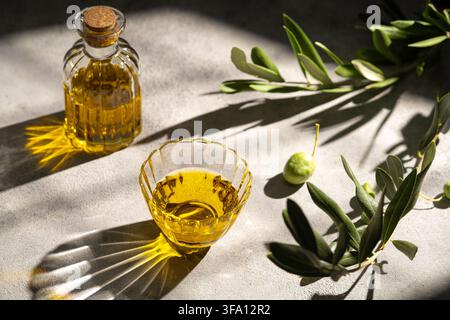 Golden olive oil in a glass bowl and corked bottle beside fresh olive branches and fruit, illuminated by dramatic sunlight and botanical shadows on a Stock Photo
