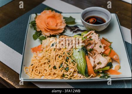 Stir-fried chicken egg noodles with fresh mixed vegetables, herbs and savory sauce, elegantly served on a square plate in a traditional Vietnamese res Stock Photo