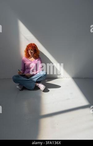 Red-haired woman sits cross-legged on a concrete floor, writing in a notebook beside a gray wall as geometric sunlight and deep shadows create a quiet Stock Photo