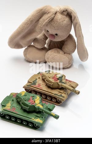 A plush bunny sits behind two camouflage toy tanks on a white background. Stock Photo