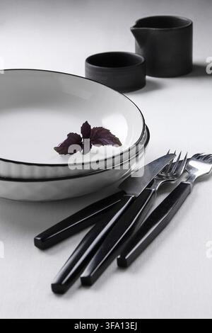 Minimalist culinary still life of black and white tableware, including plates, forks, knife and dark basil garnish, arranged on a clean light surface. Stock Photo