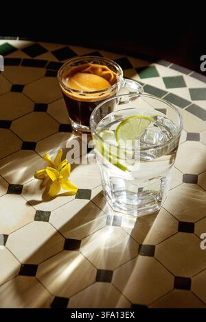 Sunlit minimalist still life of espresso and lime water with ice in clear glasses on patterned tile, accented by a yellow flower and dramatic shadows. Stock Photo