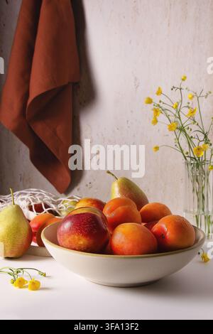Minimalist food still life of ripe peach, apricot and pear in a ceramic bowl, arranged beside yellow wildflowers and a rust-colored linen cloth agains Stock Photo
