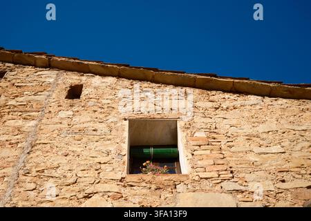 Low-angle view of a weathered stone house facade with a small flower-filled window beneath a deep blue sky in Mura, a traditional rural village in Cat Stock Photo