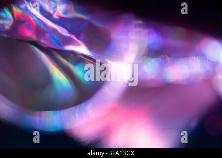 Close-up of iridescent holographic fabric reflecting vivid pink, purple, blue and green light, creating a dreamy abstract background with soft focus a Stock Photo