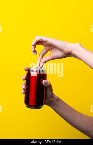 Two female hands hold and open a red aluminum beverage can against a vivid yellow background, creating a colorful studio concept for refreshment, soda Stock Photo