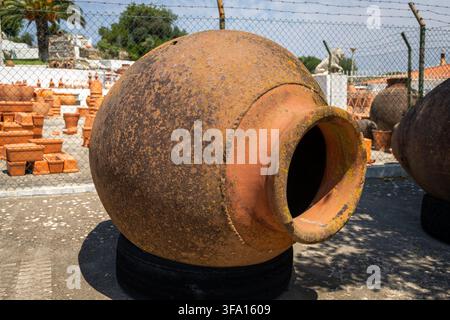 A large, old, and weathered terracotta amphora is displayed outdoors, possibly at a market or garden center, with other pottery in the background Stock Photo