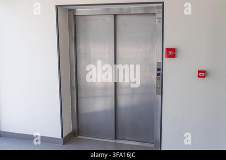 A contemporary elevator entrance featuring brushed stainless steel doors and a sleek control panel with illuminated buttons, alongside red fire alarm Stock Photo