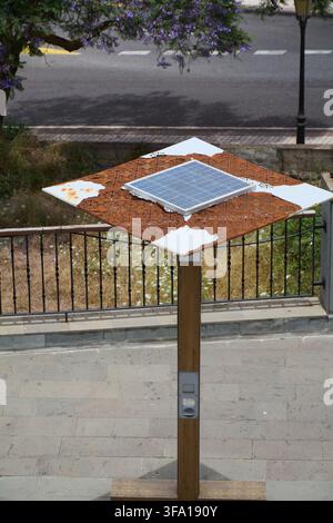Public eco-friendly solar charging station featuring a small photovoltaic panel and integrated interface on a wooden post in a street setting in Gran Stock Photo