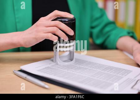 Hand pressing self inking stamp onto paperwork on desk with blurred office background, close view of document approval, office administration, record Stock Photo