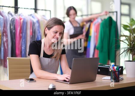 Smiling boutique owner using laptop at desk while managing clothing racks in bright fashion studio with coworker sorting colorful garments in Stock Photo