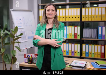 Confident businesswoman standing with folded arms in modern office beside presentation board and colorful file shelves, wearing green blazer, with Stock Photo