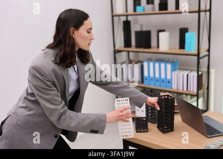 Young professional woman arranging architectural building models on desk beside laptop in modern office workspace, focusing on design planning Stock Photo