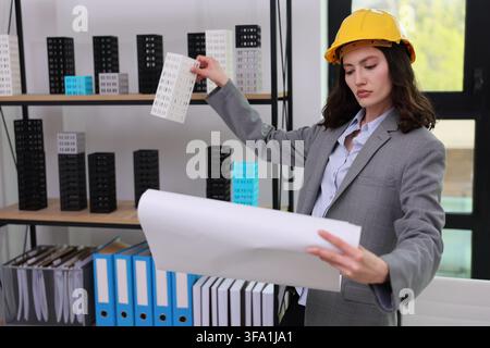 Female architect in yellow hard hat reviewing building model and blueprint in modern office with shelf of miniature towers, focused professional Stock Photo