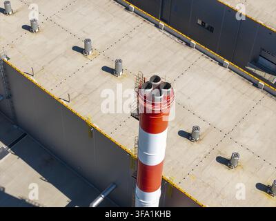 Aerial view of a red and white industrial chimney rising from a large factory roof with ventilation units in Poland. Stock Photo