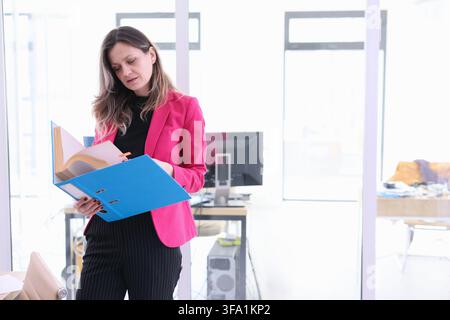 Businesswoman in pink blazer reviewing documents in bright modern office while holding blue folder and standing near computer workstation, focused on Stock Photo